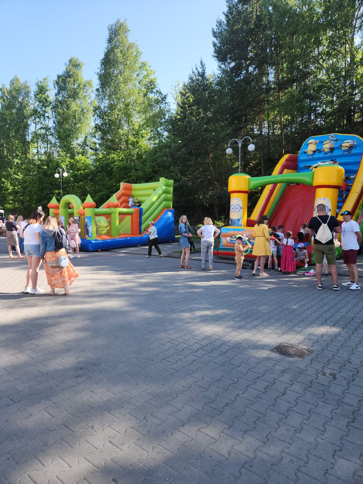 Dwa dmuchane zamki z motywami bajkowymi, otoczone grupą dzieci i dorosłych na placu zabaw z kostki brukowej, w tle drzewa i latarnie.