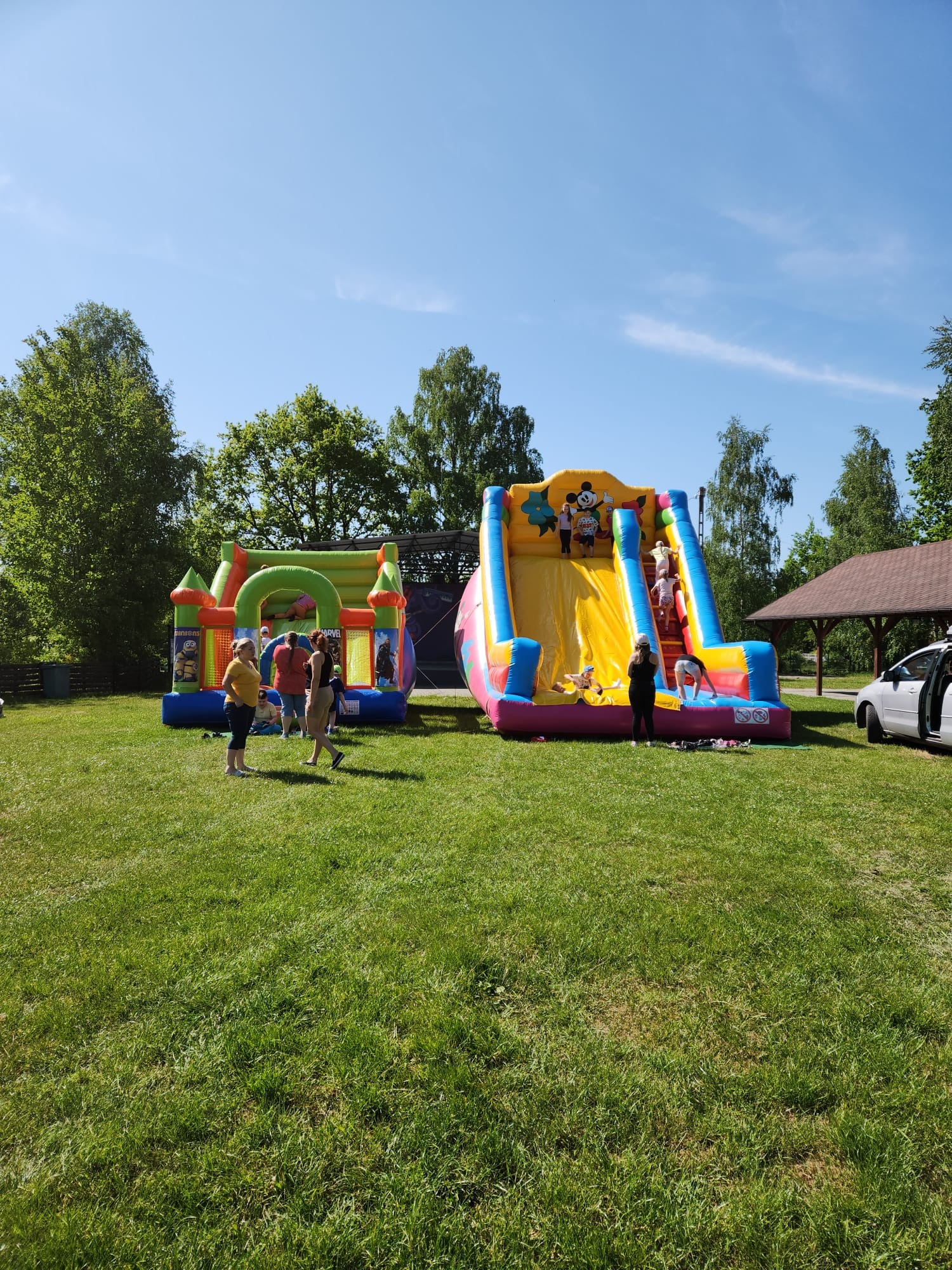 Dwa dmuchane zamki na zielonej trawie, jeden z motywem Minionków i Marvela, drugi z dużą żółtą zjeżdżalnią ozdobioną postaciami z bajek, dzieci korzystające z atrakcji, w tle drzewa i błękitne niebo.