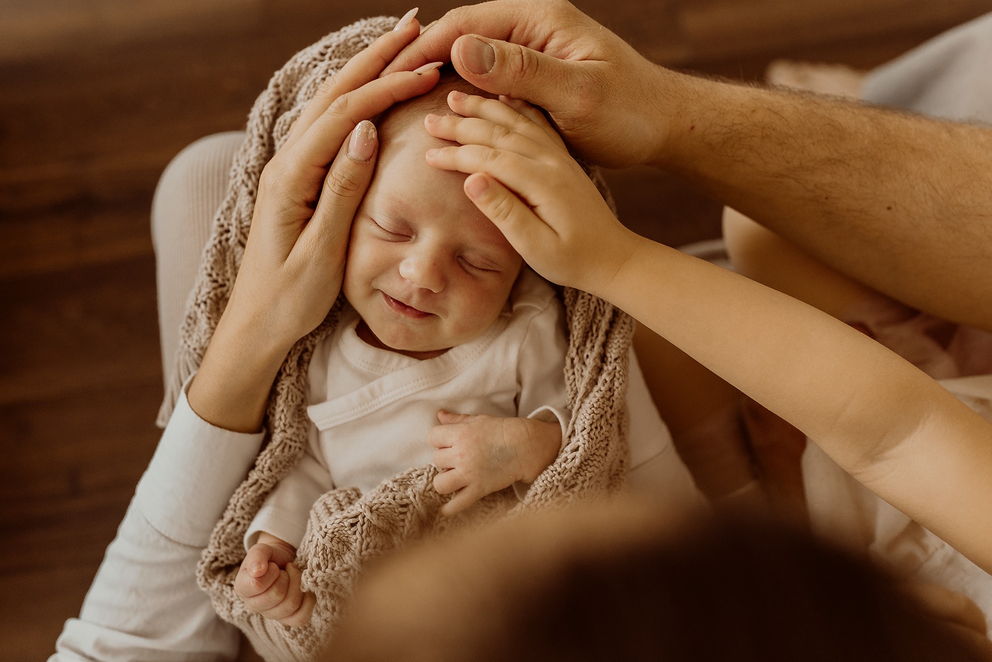 Śpiący noworodek otulony beżowym kocykiem, dłonie rodziców delikatnie dotykają jego głowy, zarys dłoni starszego dziecka.