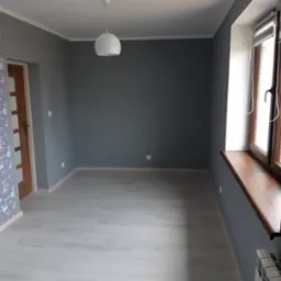 Pomalowane na szaro wnętrze pokoju z jasnymi panelami podłogowymi, drewnianą stolarką okienną i wzorzystą tapetą na fragmencie ściany przy drzwiach.