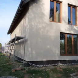 Świeżo otynkowana ściana domu jednorodzinnego z drewnianymi oknami, widoczne rusztowanie, teren wokół budynku w trakcie porządkowania, pozostałości materiałów budowlanych.