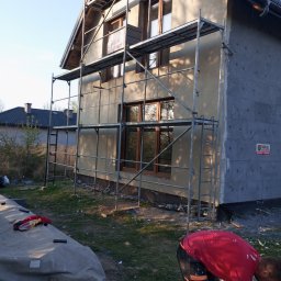Fasady elewacje - Dom w trakcie ocieplania, widoczne rusztowania, częściowo otynkowana elewacja, ocieplenie styropianem na ścianie bocznej, pracownik przygotowujący zaprawę.