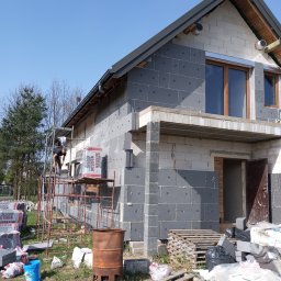 Fasady elewacje - Dom w trakcie docieplania elewacji styropianem grafitowym, widoczne rusztowanie, palety z materiałem izolacyjnym i pracownik na rusztowaniu w słoneczny dzień.