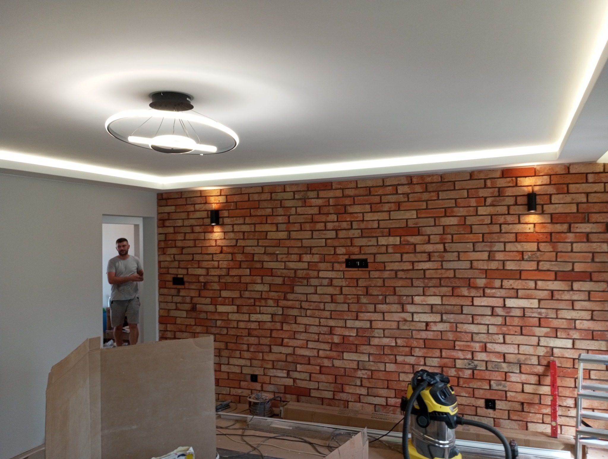 Pokój z ceglaną ścianą, oświetleniem LED pod sufitem i nowoczesną lampą wiszącą, widoczny mężczyzna w tle, częściowo zasłonięte elementy wyposażenia i odkurzacz na pierwszym planie.