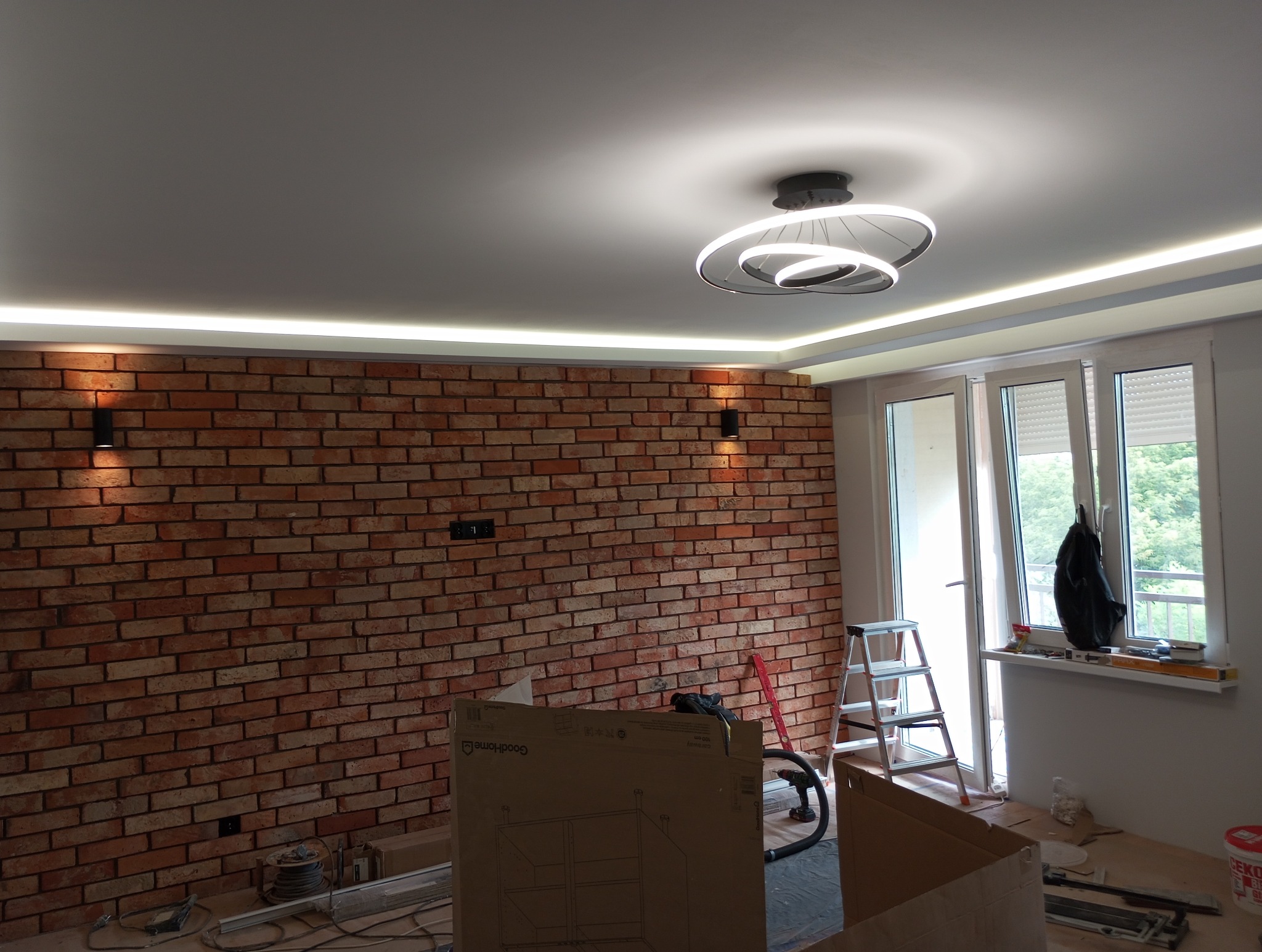 Ściana z cegły klinkierowej z oświetleniem punktowym i ledowym, nowoczesna lampa sufitowa, okno balkonowe z roletami, drabina, karton po meblach i narzędzia na podłodze w trakcie remontu.