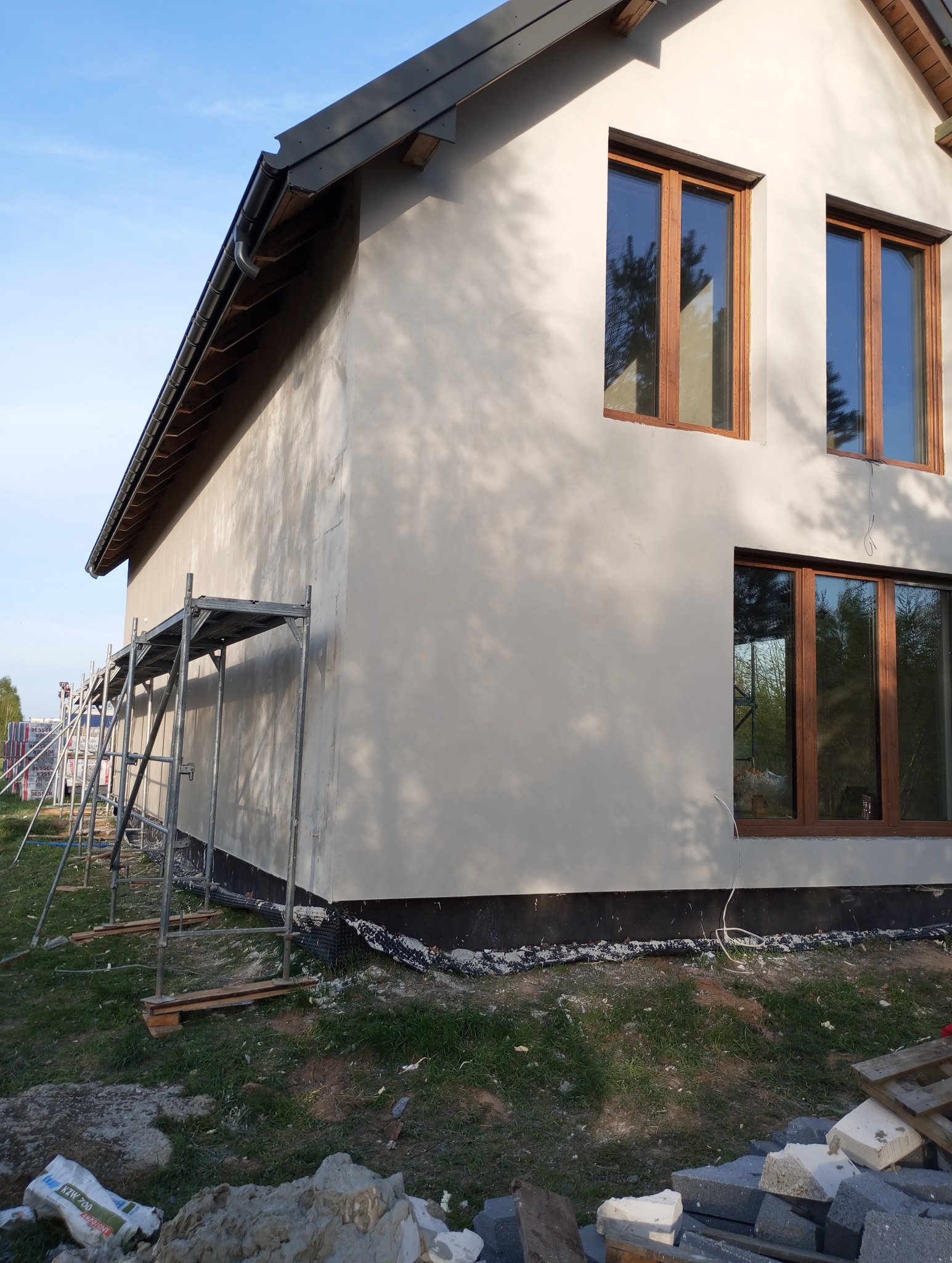 Świeżo otynkowana ściana domu jednorodzinnego z drewnianymi oknami, widoczne rusztowanie, teren wokół budynku w trakcie porządkowania, pozostałości materiałów budowlanych.
