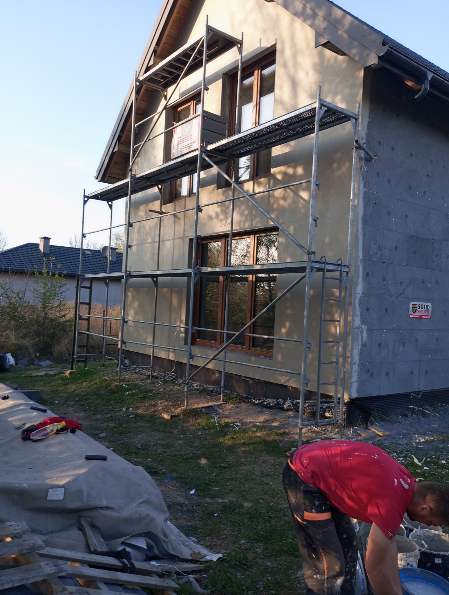 Dom w trakcie ocieplania, widoczne rusztowania, częściowo otynkowana elewacja, ocieplenie styropianem na ścianie bocznej, pracownik przygotowujący zaprawę.