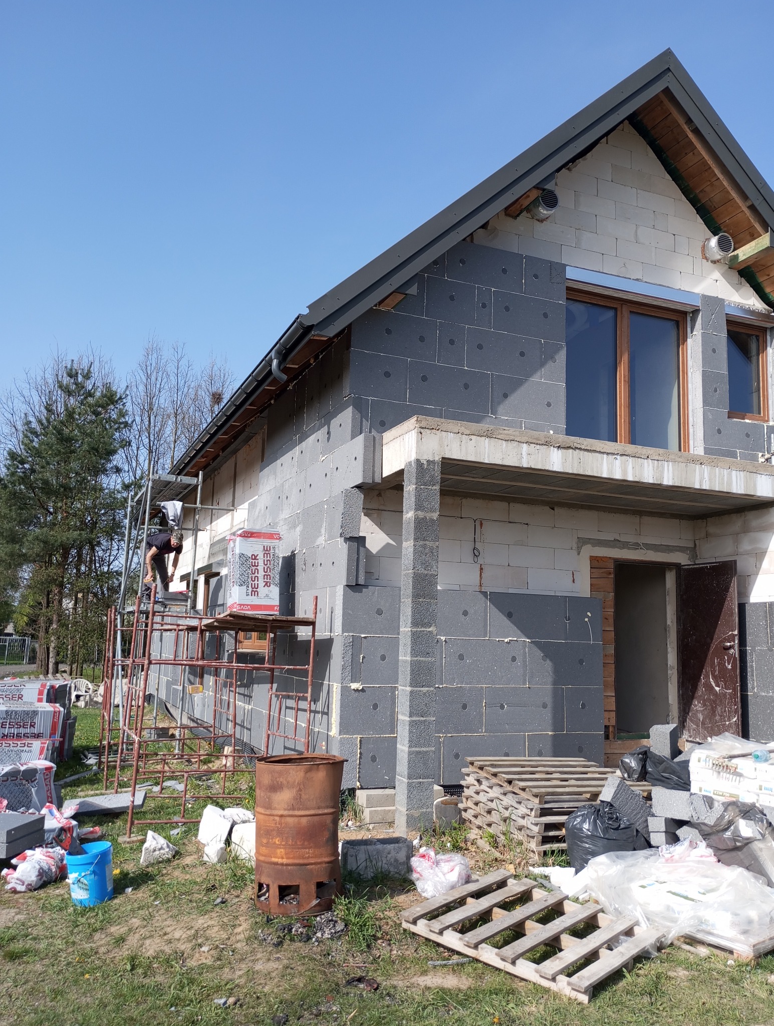 Dom w trakcie docieplania elewacji styropianem grafitowym, widoczne rusztowanie, palety z materiałem izolacyjnym i pracownik na rusztowaniu w słoneczny dzień.
