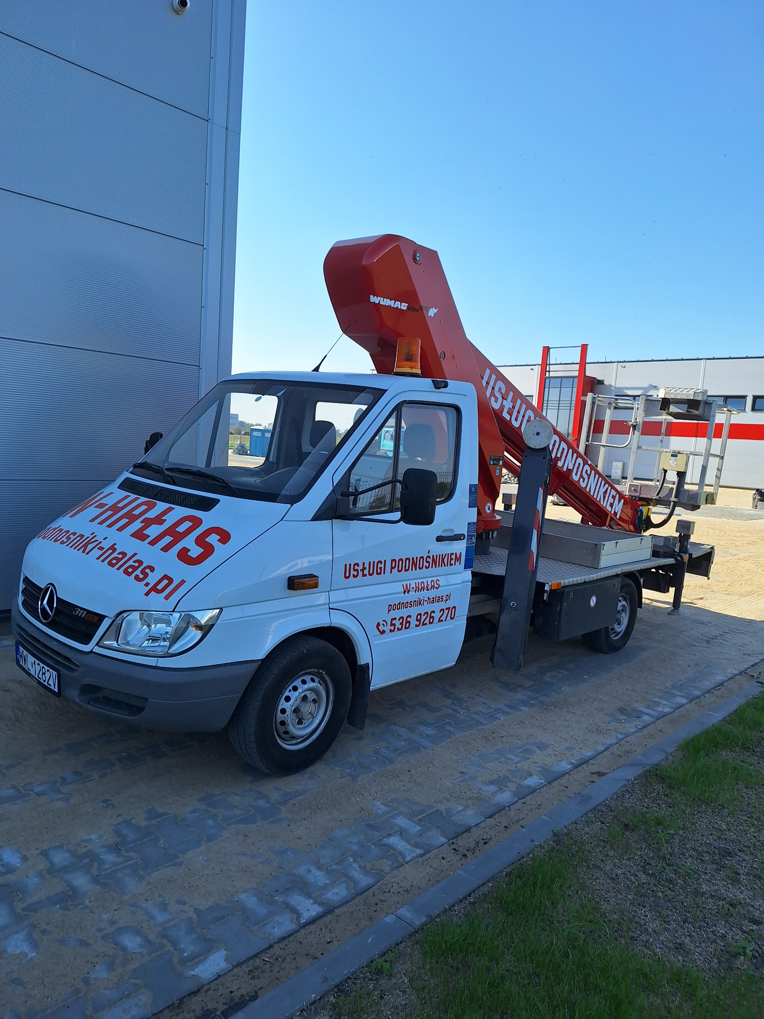 Biały samochód dostawczy z czerwonym wysięgnikiem koszowym WUMAG Elephant zaparkowany na kostce brukowej przed budynkiem przemysłowym.