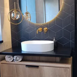 Nowoczesna łazienka z okrągłym lustrem podświetlonym LED, umywalką nablatową, złotą armaturą i geometrycznymi płytkami na ścianie.