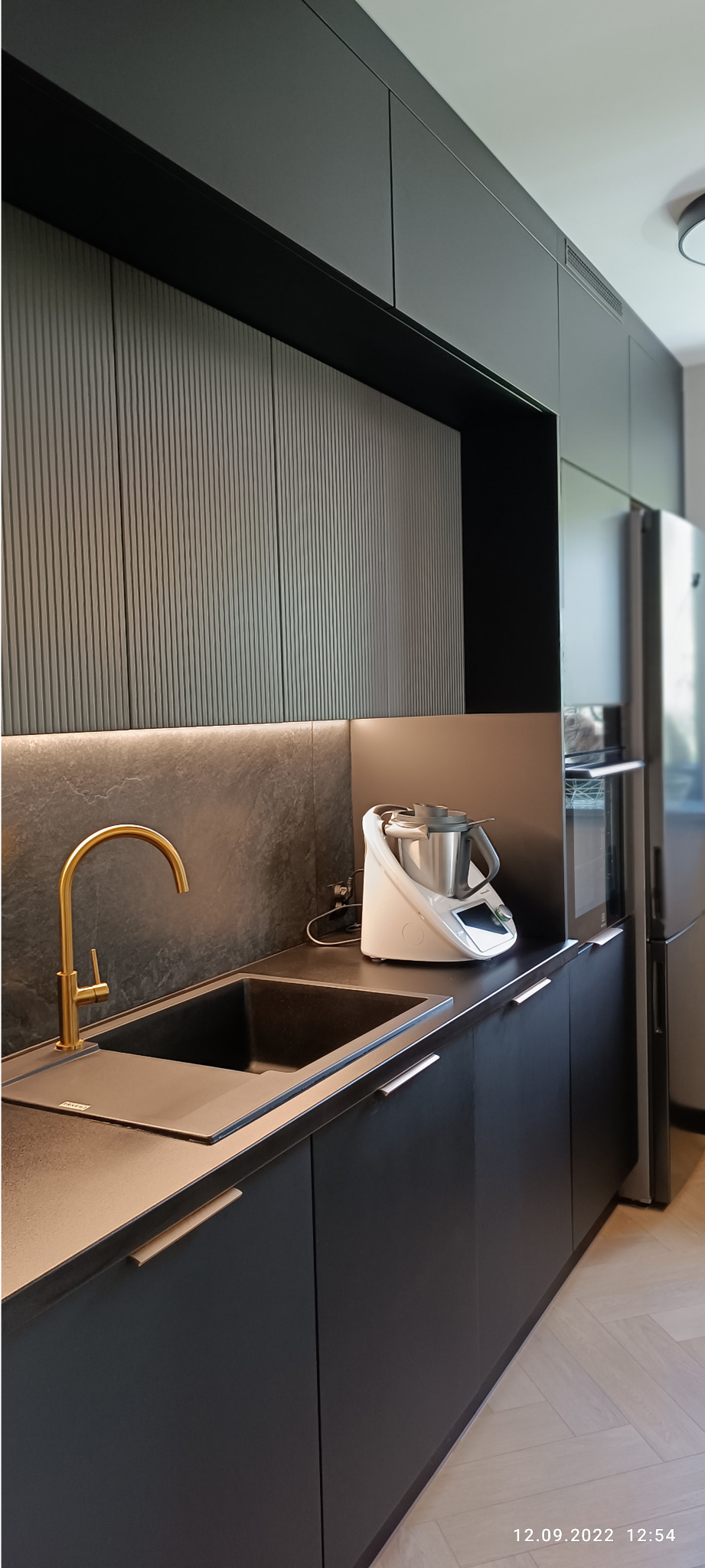 Nowoczesna, czarna kuchnia z matowymi frontami, złotą baterią i ciemnym blatem, oświetlenie LED pod szafkami, widoczny robot kuchenny.