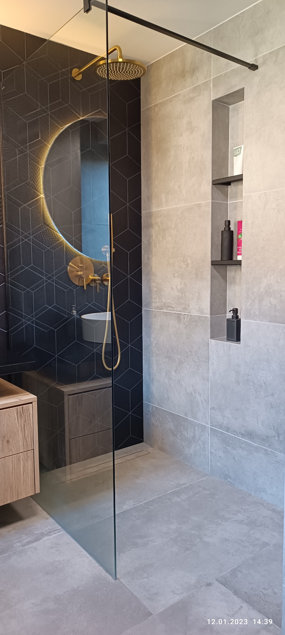 Nowoczesna łazienka z czarnymi heksagonalnymi płytkami, okrągłym lustrem z podświetleniem LED, złotą armaturą prysznicową i szarą płytką imitującą beton, z wnęką na kosmetyki.