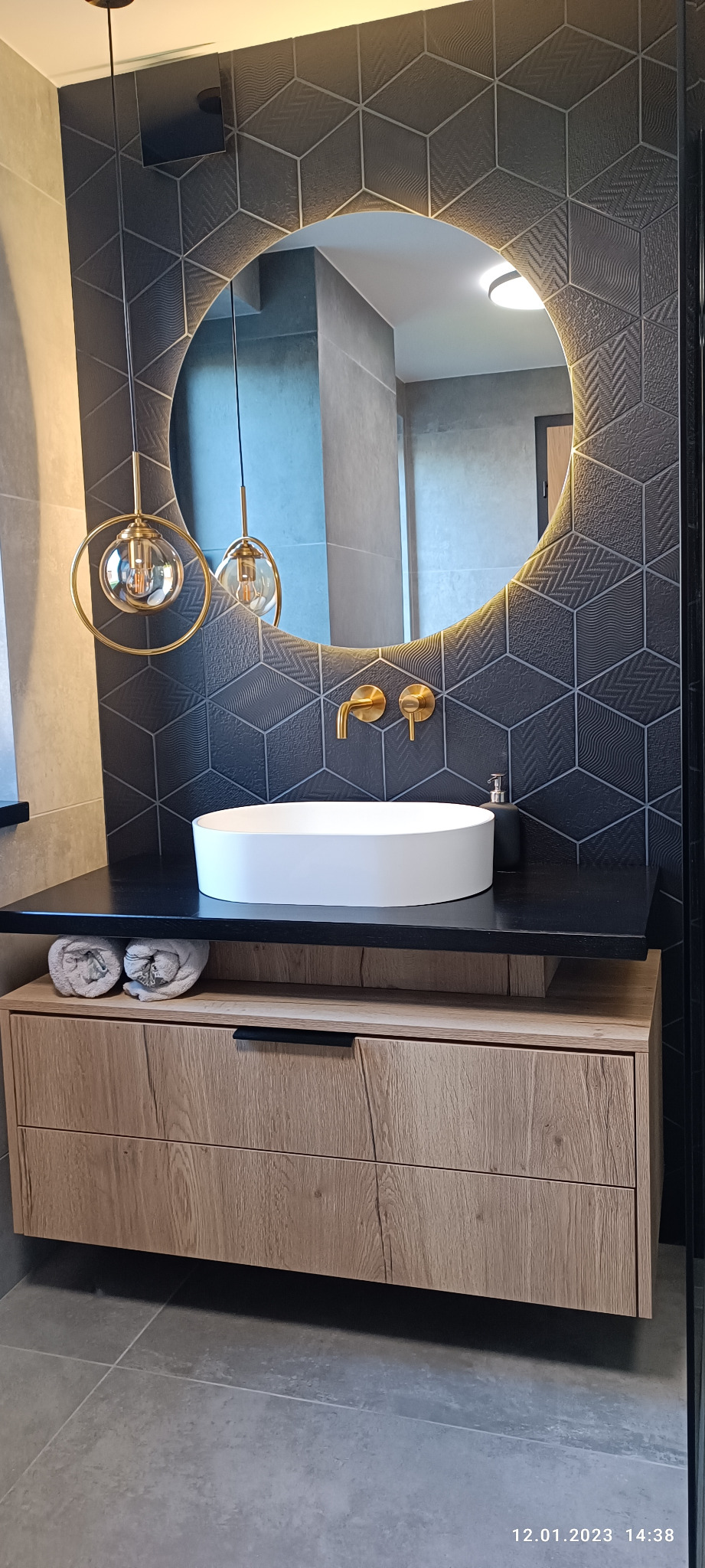 Nowoczesna łazienka z okrągłym lustrem podświetlonym LED, umywalką nablatową, złotą armaturą i geometrycznymi płytkami na ścianie.
