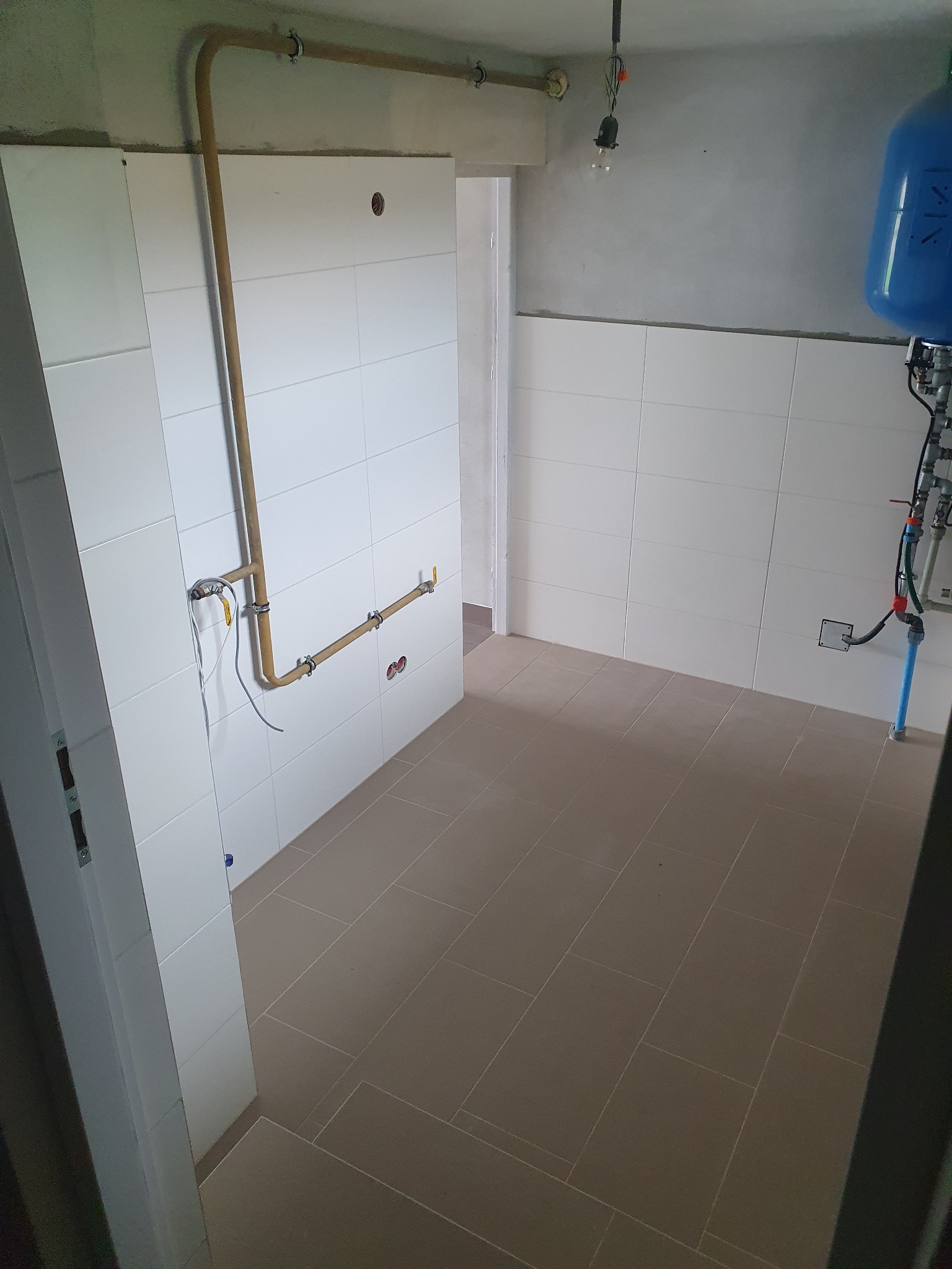 Wnętrze pomieszczenia w trakcie prac wykończeniowych: białe płytki na ścianach, brązowe płytki podłogowe, rury instalacji wodnej na ścianie, wiszący bojler i surowy betonowy sufit z wiszącą żarówką.