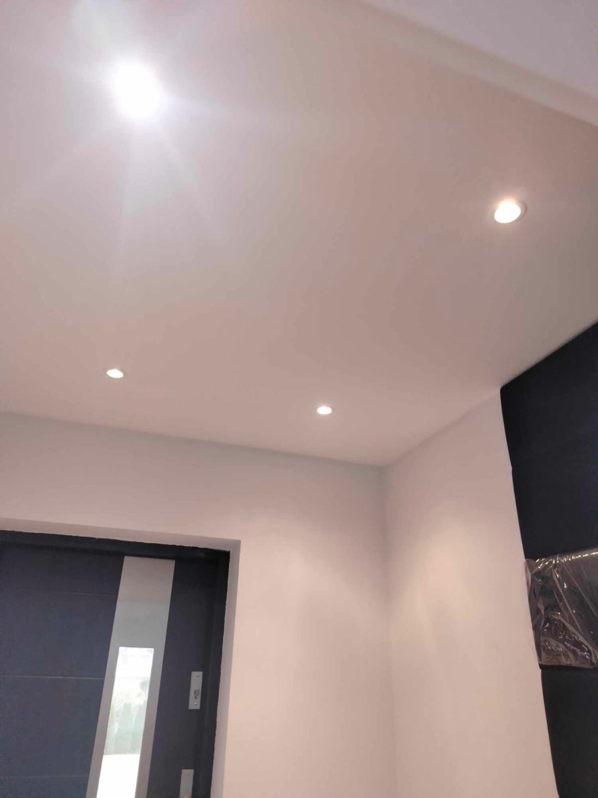 Białe wnętrze z trzema punktowymi lampami w suficie, świeżo pomalowane ściany i nowoczesne drzwi z szarą ramą.