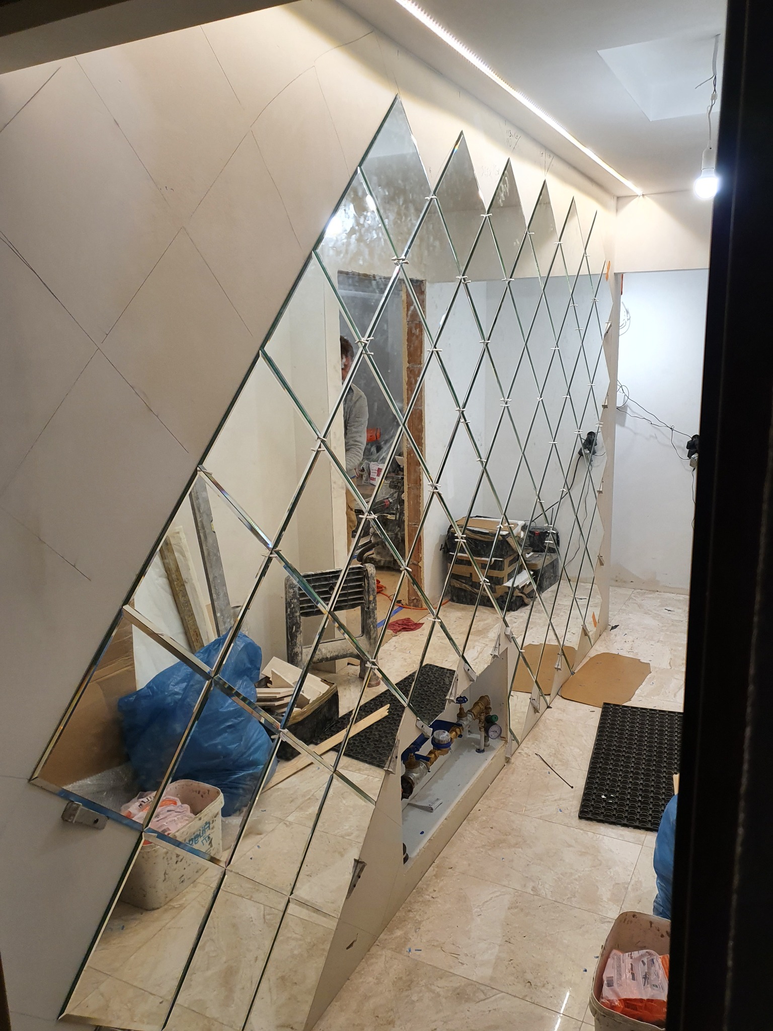 Montaż dekoracyjnych, romboidalnych luster na ścianie, widoczne przygotowane podłoże i odblask pracownika w lustrach.