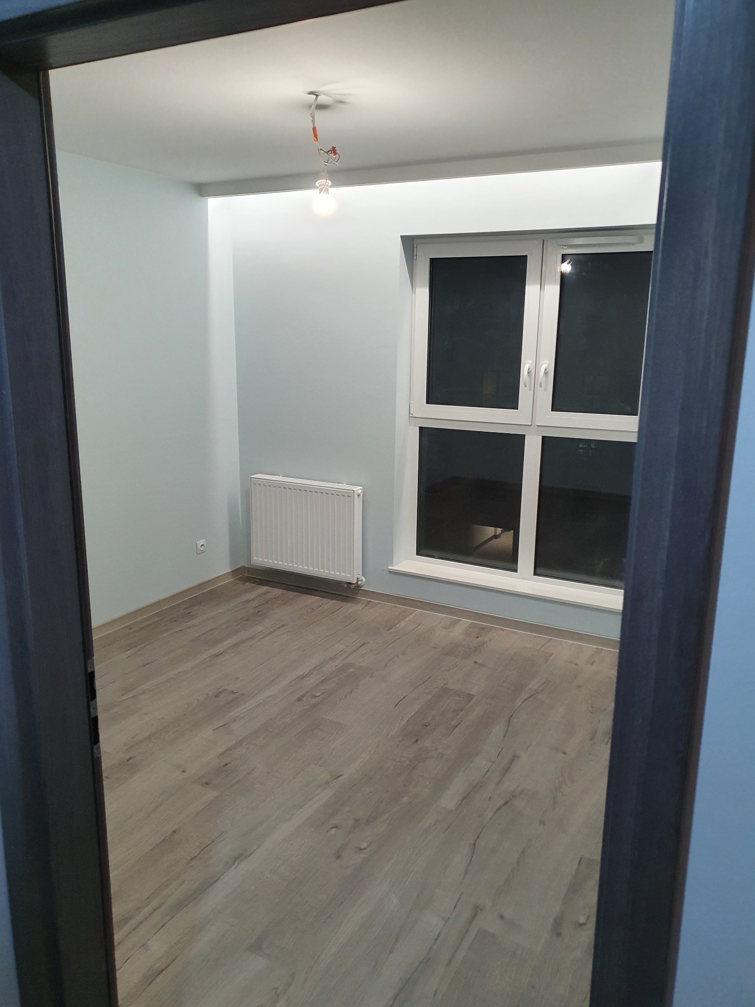 Pomalowane na jasno pomieszczenie z ułożonymi panelami podłogowymi, widok przez futrynę drzwiową, żarówka zwisająca z sufitu.