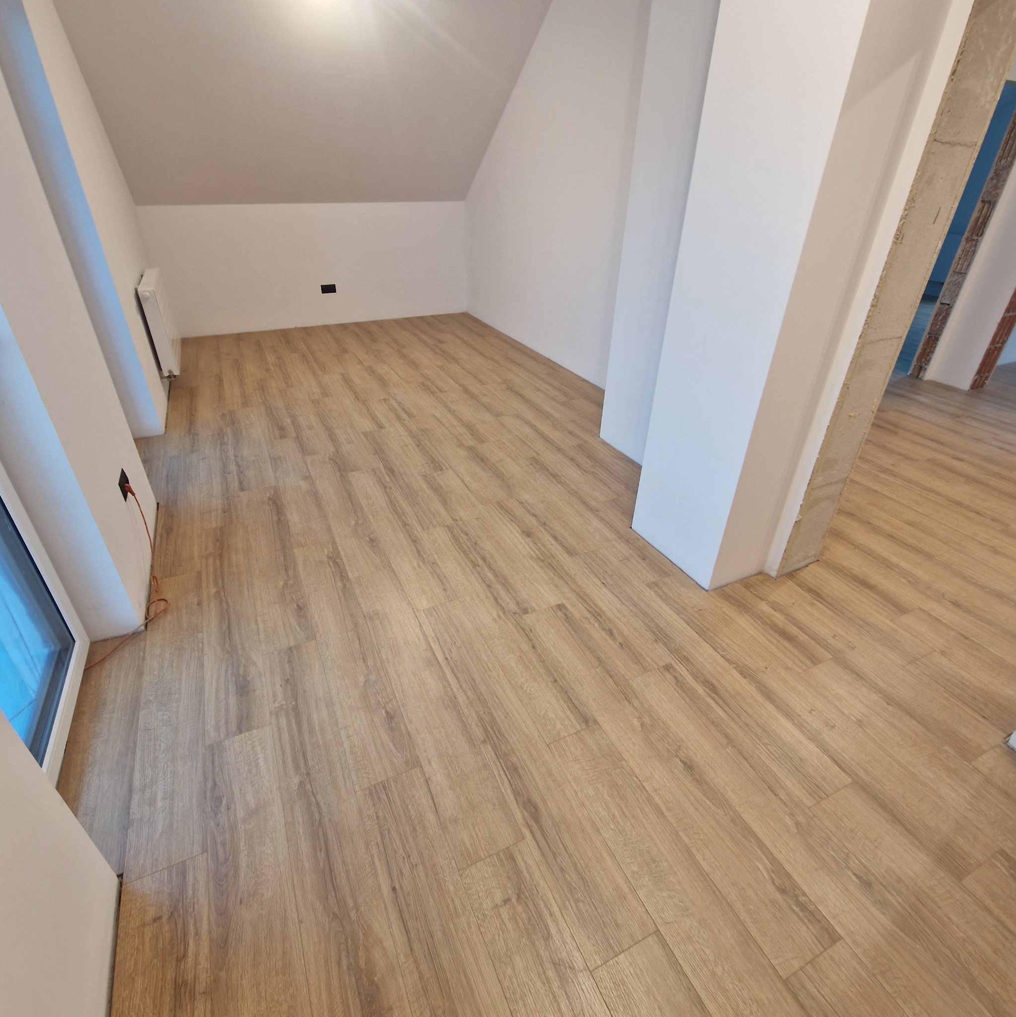 Jasne panele podłogowe ułożone w pokoju na poddaszu, widoczne białe ściany i filar. Fragment okna po lewej. Gniazdko elektryczne z kablem przy ścianie.