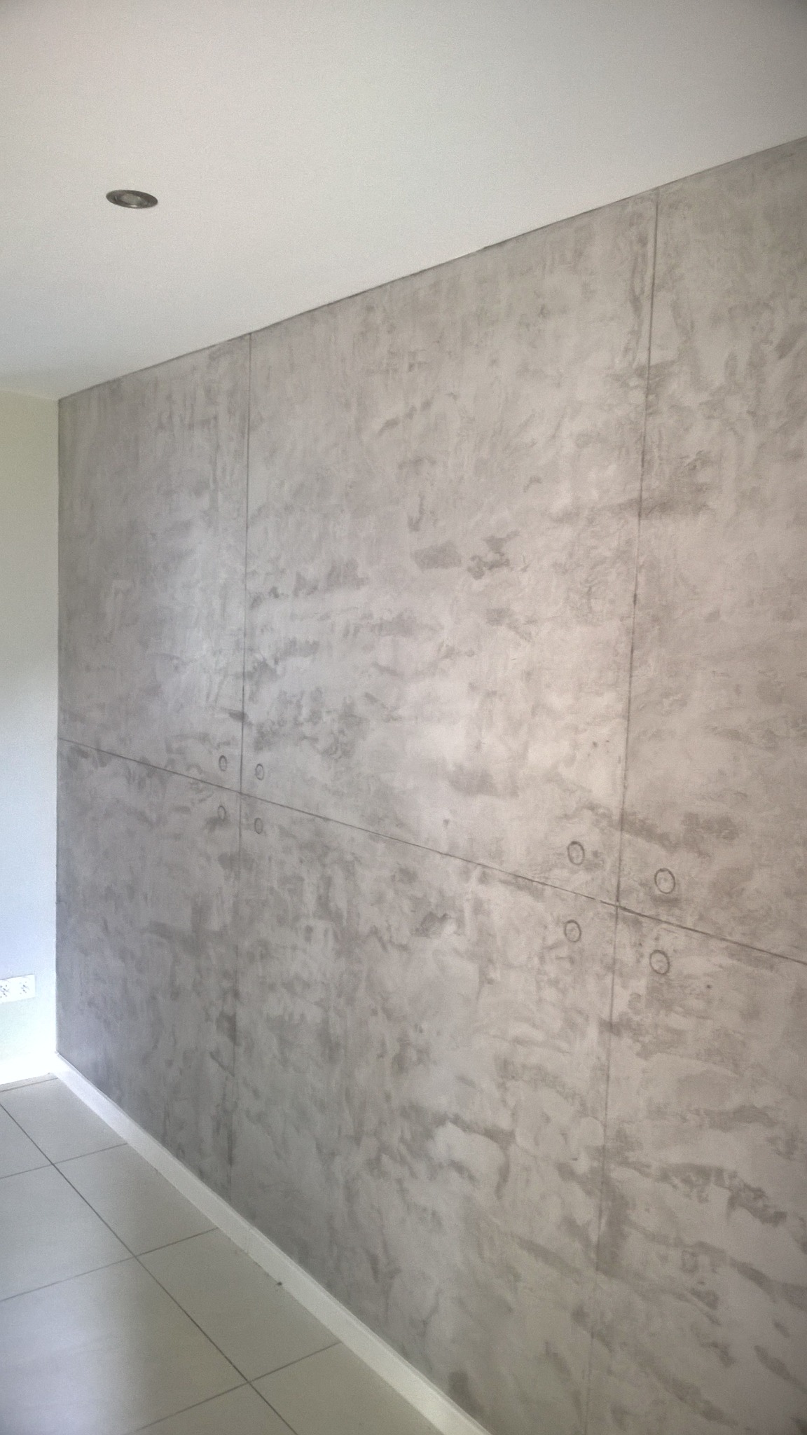 Ściana wykończona szarą, dekoracyjną gładzią imitującą beton, podzielona na panele z widocznymi otworami montażowymi.