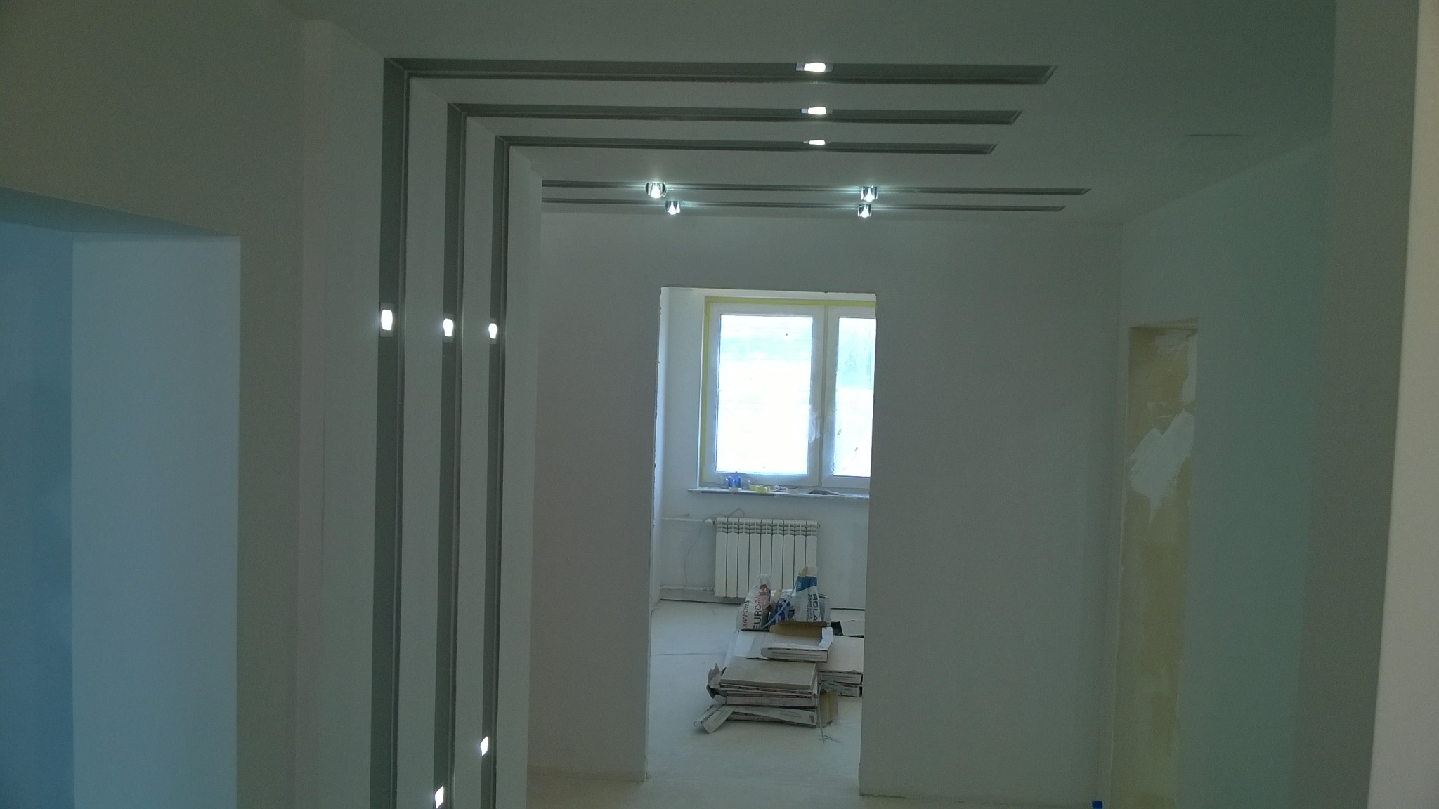Wykończone wnętrze z podwieszanym sufitem z oświetleniem punktowym i pionowymi listwami oświetleniowymi na ścianach, widok na pokój z oknem i grzejnikiem, kartony na podłodze.