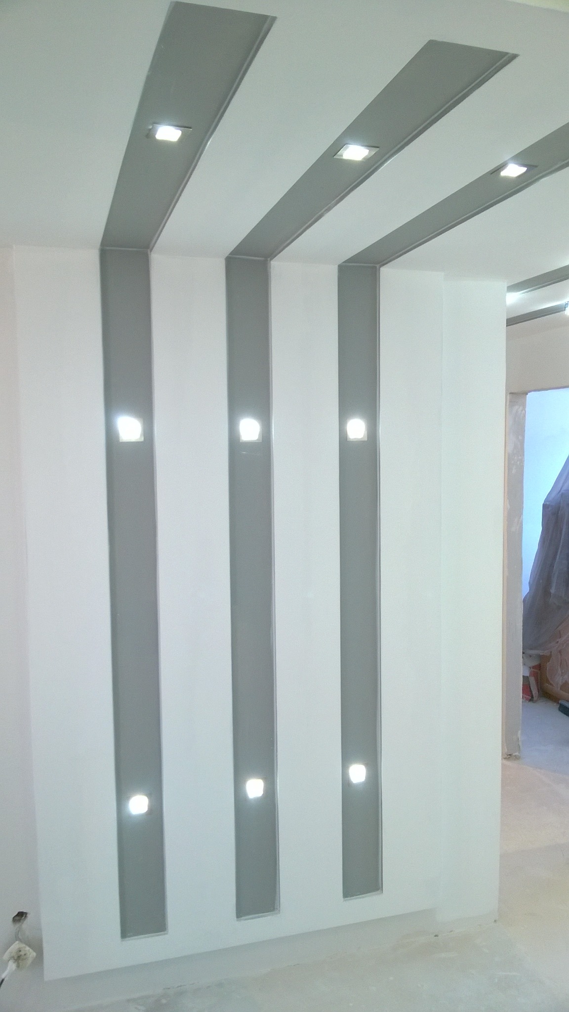 Biała ściana z szarymi pionowymi pasami i wbudowanymi punktowymi lampami LED, widok na sufit z podobnym wzorem, wnętrze w trakcie wykańczania.