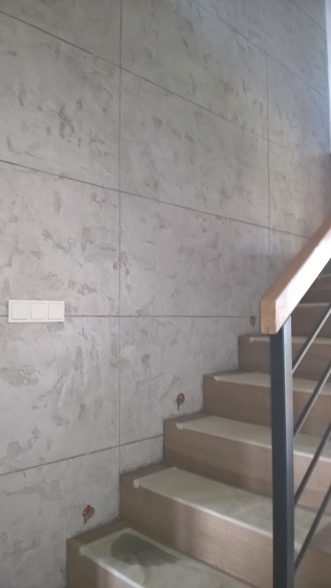 Ściana wykończona gładzią dekoracyjną w odcieniach szarości, z widocznymi schodami z drewnianą balustradą i metalowymi elementami. Na ścianie zamontowany potrójny włącznik światła.