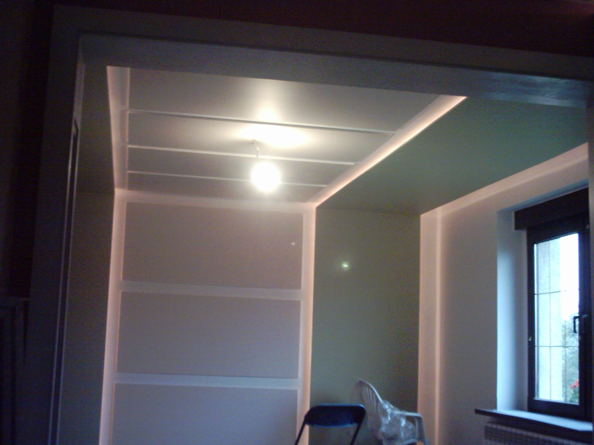 Wykończone wnętrze z podwieszanym sufitem z liniowym oświetleniem LED i centralną żarówką, widoczne ściany w odcieniach szarości i zieleni oraz okno z ciemną ramą.