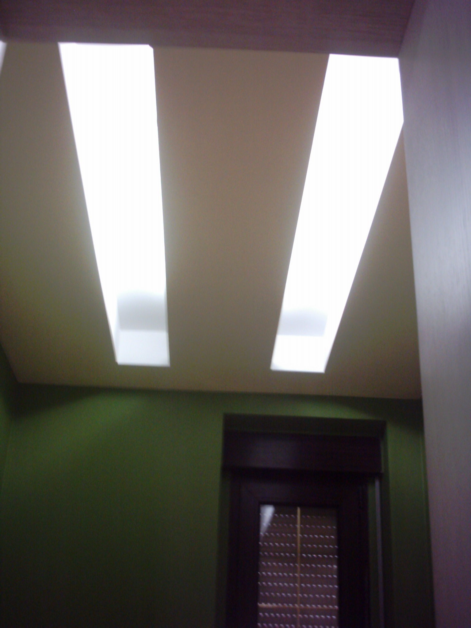 Dwa prostokątne świetliki w jasnym suficie, zielona ściana i ciemne drzwi w tle.