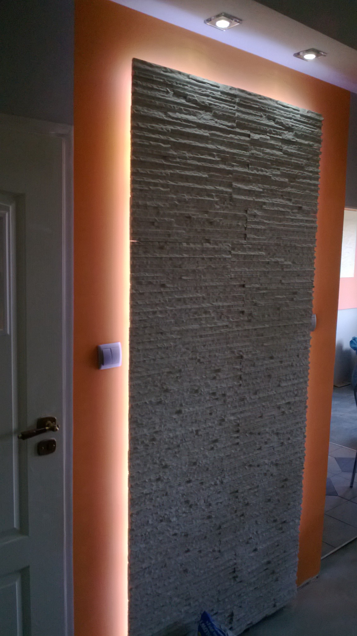 Fragment ściany z dekoracyjną okładziną imitującą kamień, podświetloną pionowym pasem ciepłego światła, sąsiadującej z pomarańczową ścianą i białą futryną drzwiową z mosiężną klamką.