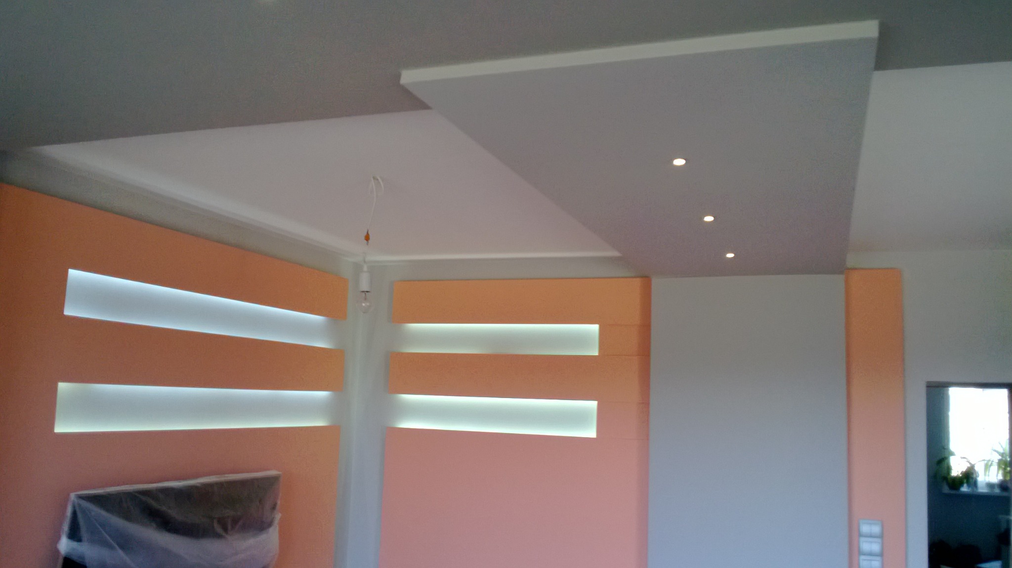 Nowoczesna aranżacja wnętrza z podwieszanym sufitem z punktowym oświetleniem i ścianą z podświetlanymi wnękami, z widocznym fragmentem telewizora pod folią ochronną.