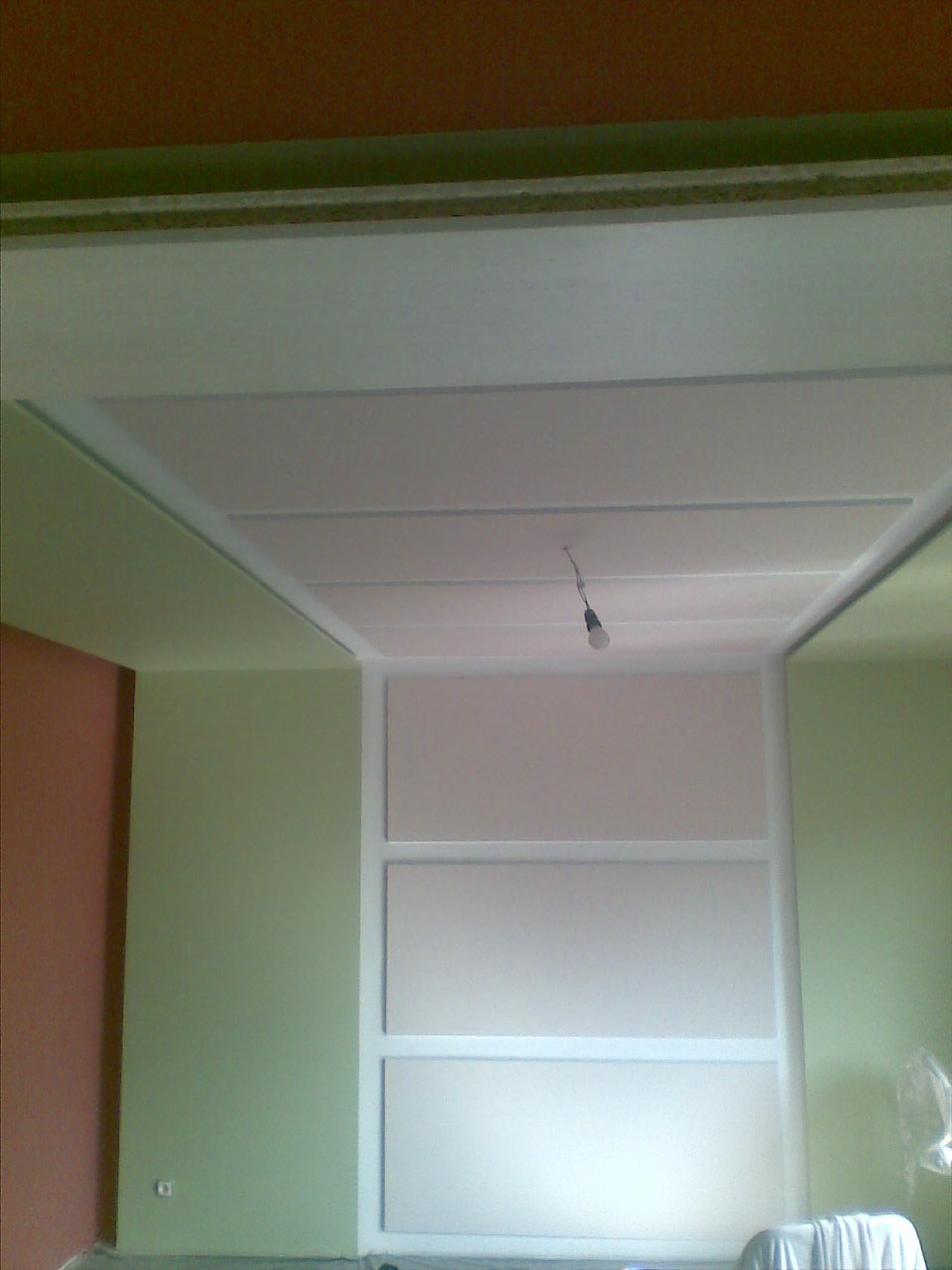 Pokój w trakcie remontu: świeżo pomalowane ściany w odcieniach zieleni i brązu, biały sufit z widoczną żarówką, ściana z wnękami w trakcie wykańczania.