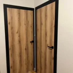Dwa skrzydła drzwiowe wewnętrzne z okleiną imitującą drewno i czarną ramą, widoczne z perspektywy podłogi z parkietu.