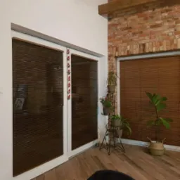 Wnętrze z drewnianą podłogą, białymi ścianami i ceglaną ścianą z oknem przesłoniętym brązowymi żaluzjami poziomymi; dwuskrzydłowe drzwi z podobnymi żaluzjami, ozdobione świątecznymi zawieszkami...