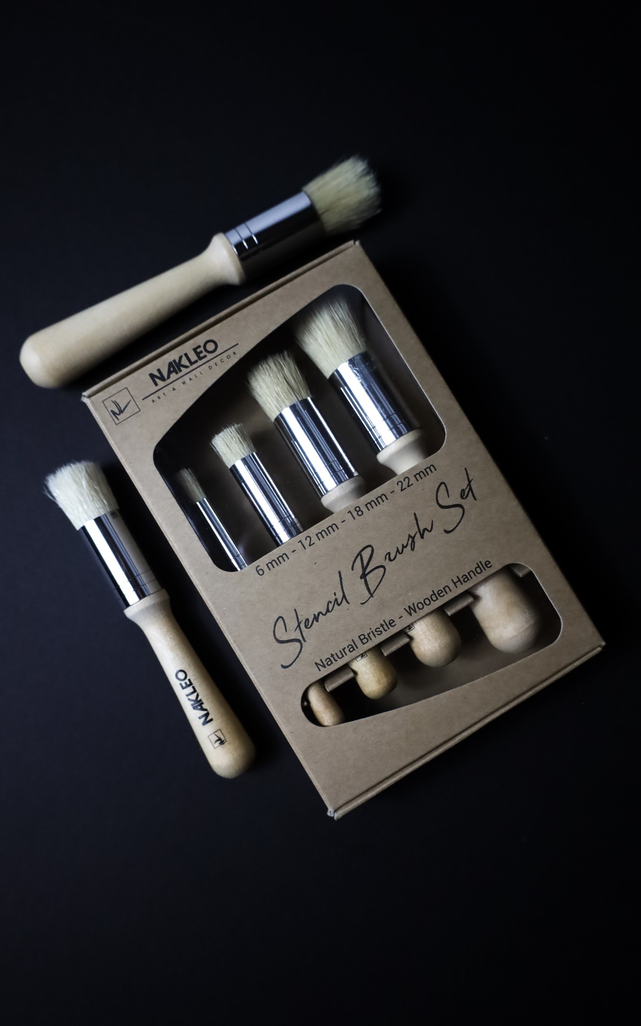 Zestaw pędzli do szablonów Nakleo z naturalnego włosia i drewnianymi uchwytami, w oryginalnym kartonowym opakowaniu na czarnym tle. Jeden pędzel leży obok opakowania, eksponując logo firmy.