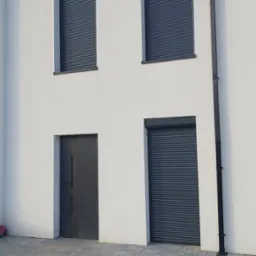 Nowoczesny dom z białymi ścianami, szarą stolarką okienną i drzwiową oraz trzema roletami zewnętrznymi w kolorze antracytowym.
