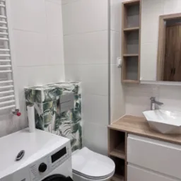 Nowoczesna łazienka z białą armaturą, pralką z przodu i toaletą z motywem liści, umywalka nablatowa o geometrycznym kształcie, podłoga z paneli imitujących drewno, białe płytki na ścianach,...