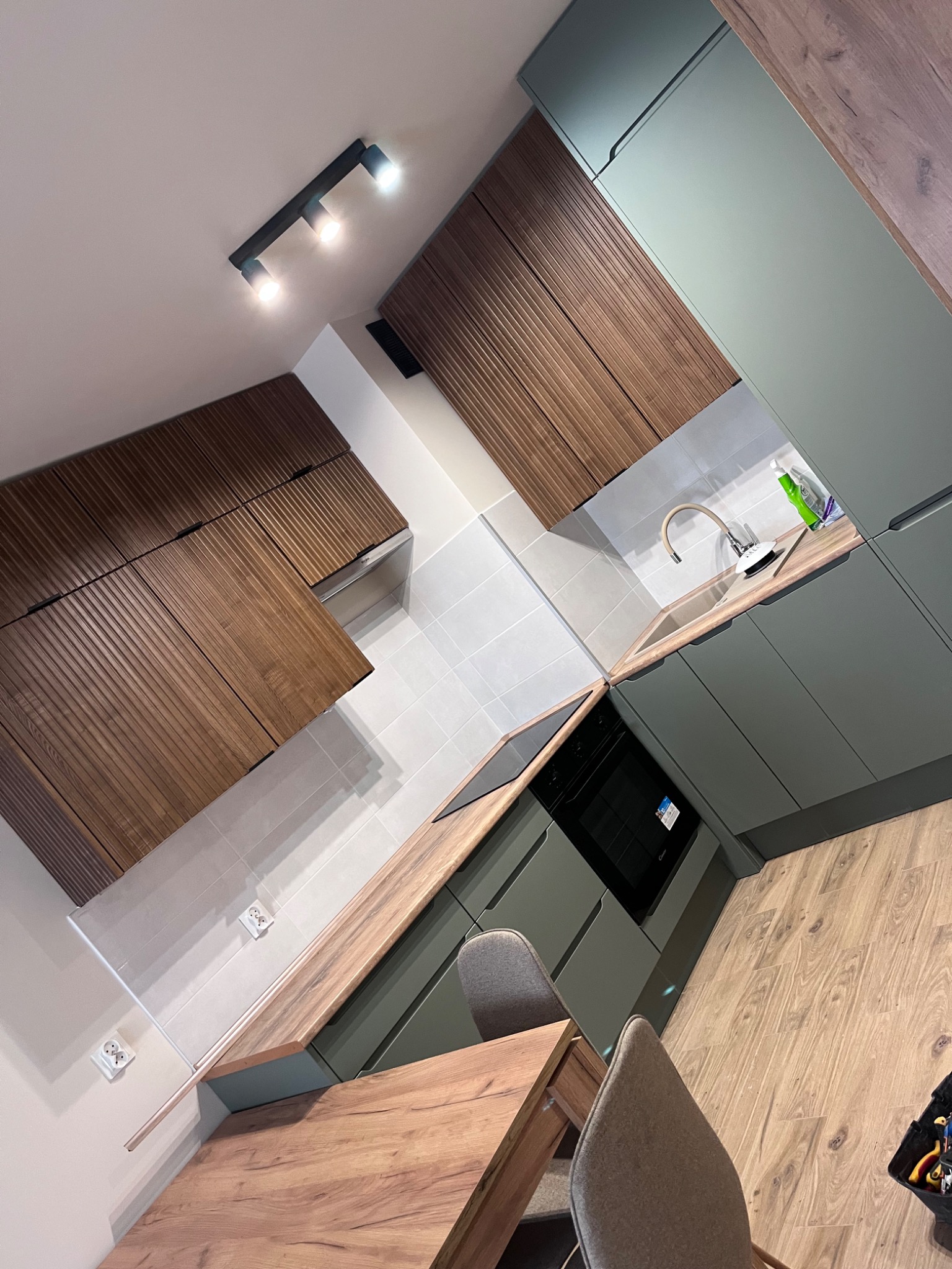 Nowoczesna kuchnia z drewnianymi frontami górnych szafek i zielonymi dolnymi szafkami, z blatem imitującym drewno i szarymi płytkami na ścianie, oświetlona trzema punktowymi lampami sufitowymi,...