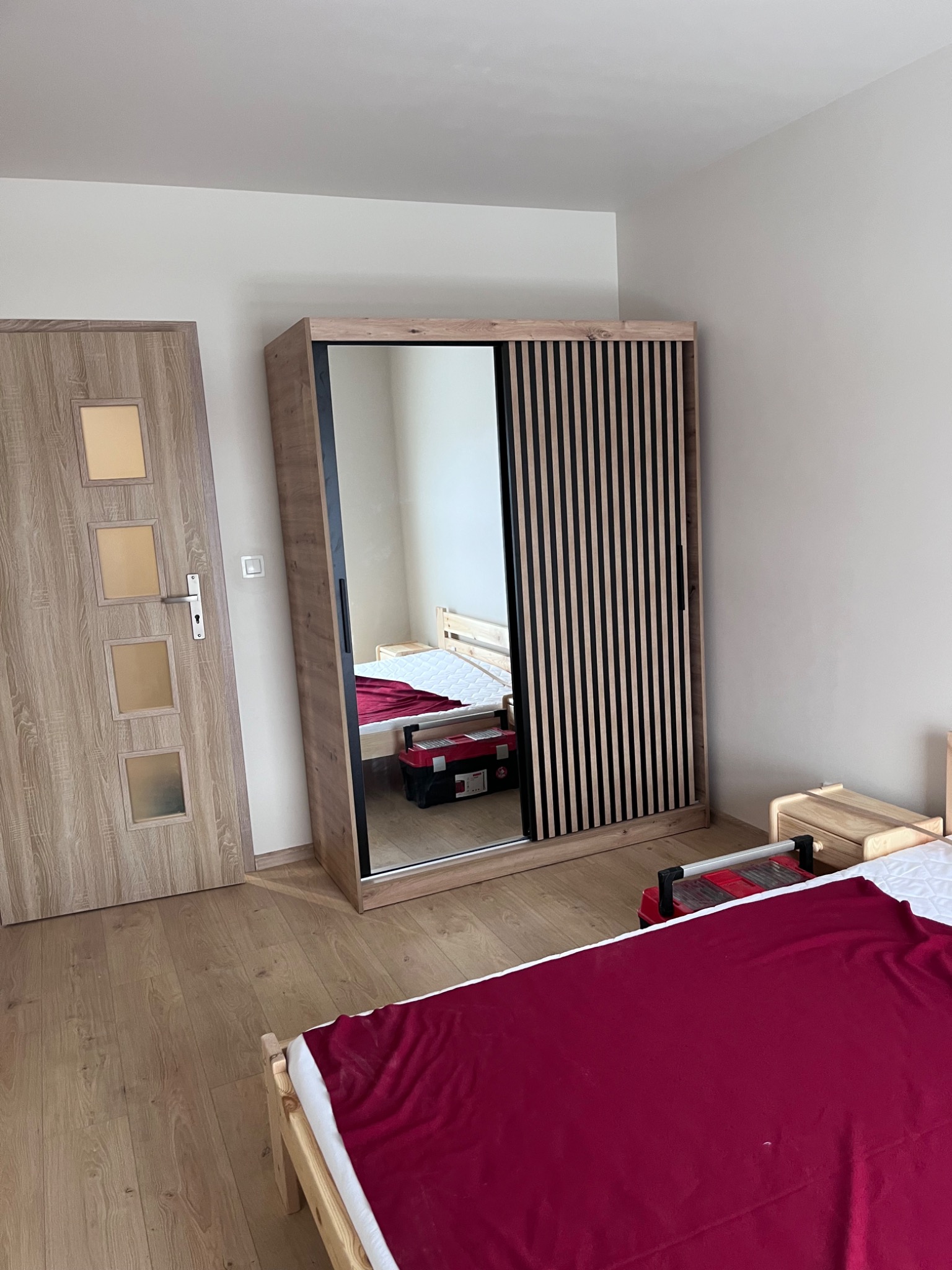 Wnętrze pokoju z drewnianą podłogą, szafa przesuwna z lustrem i dekoracyjnymi listwami, widoczna skrzynka z narzędziami, łóżko z bordową narzutą, drzwi z przeszkleniami.
