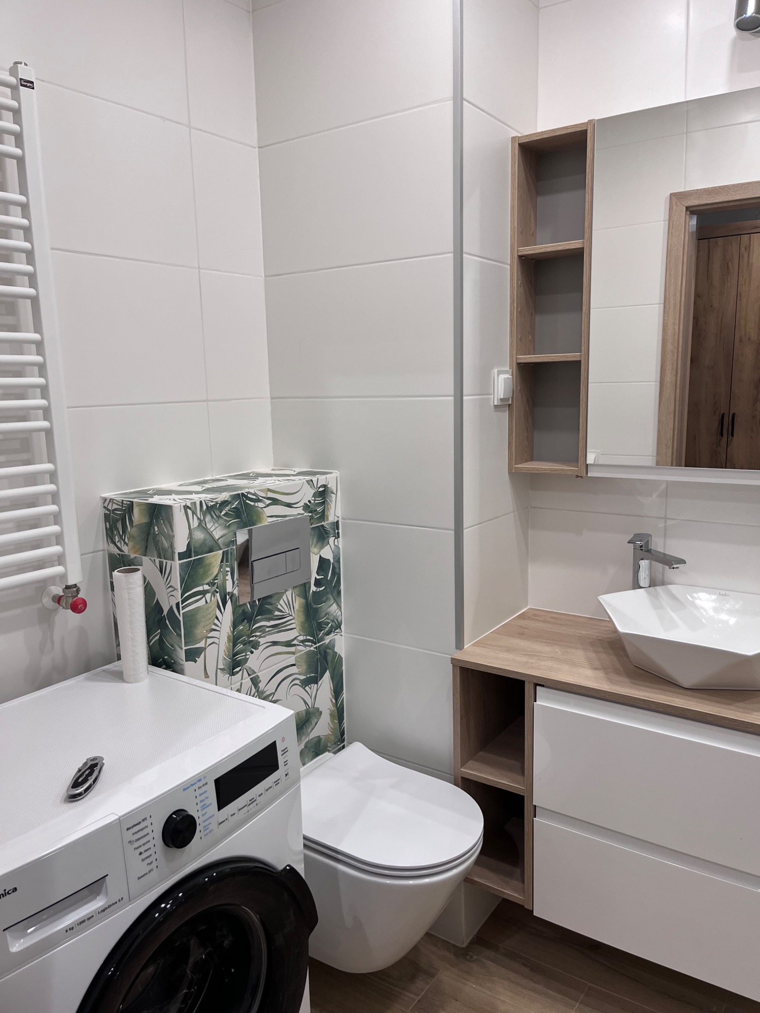 Nowoczesna łazienka z białą armaturą, pralką z przodu i toaletą z motywem liści, umywalka nablatowa o geometrycznym kształcie, podłoga z paneli imitujących drewno, białe płytki na ścianach,...