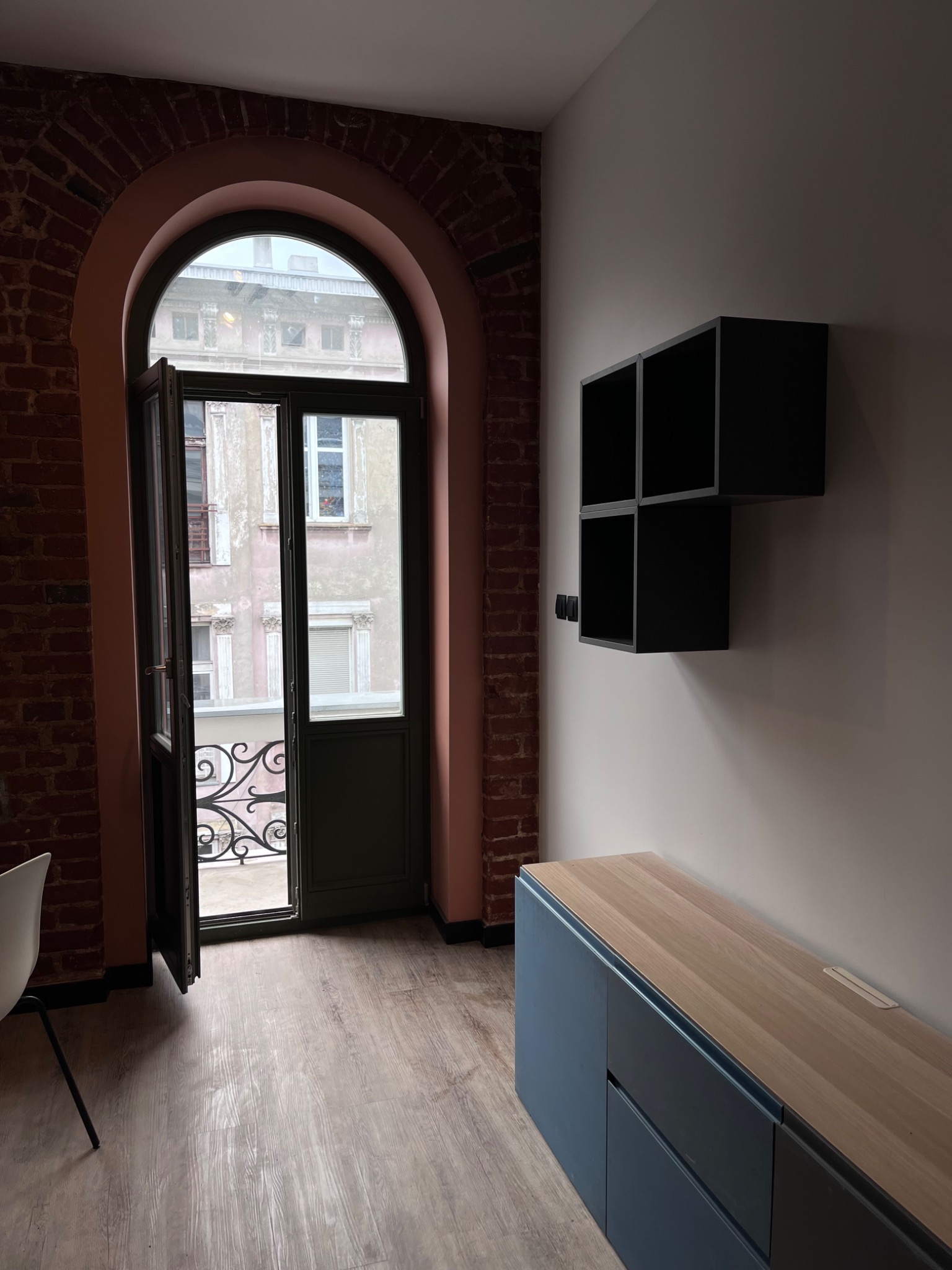 Wnętrze pokoju z odnowioną ceglaną ścianą, otwartym balkonem z kutą balustradą, drewnianą podłogą i minimalistycznymi czarnymi półkami na białej ścianie.