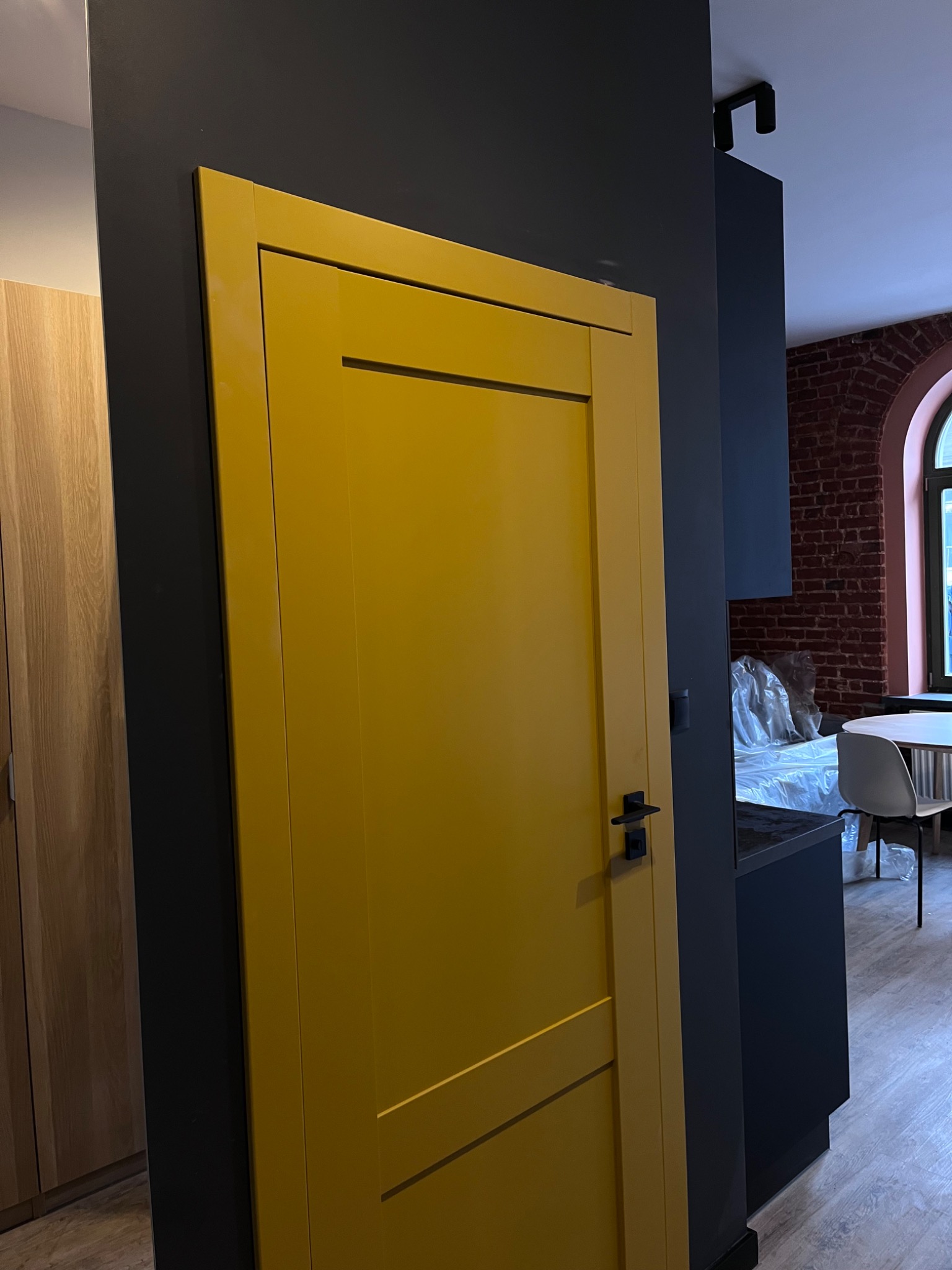 Żółte drzwi wewnętrzne z czarną klamką, zamontowane w nowocześnie wykończonym wnętrzu z ciemnymi ścianami i widoczną częścią kuchni z ceglaną ścianą w tle.