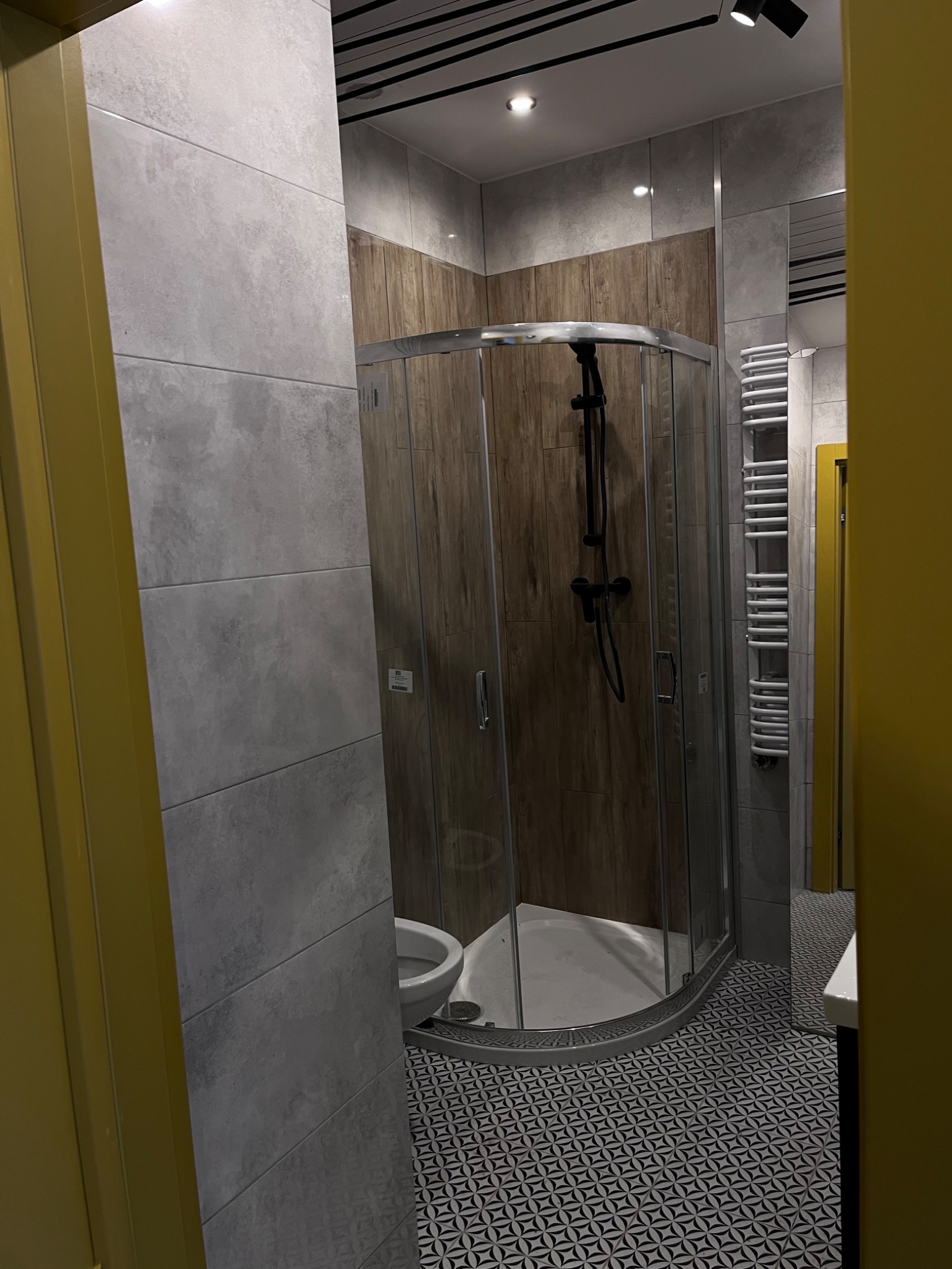 Nowoczesna łazienka z kabiną prysznicową typu walk-in z drewnopodobnymi panelami, szare płytki wielkoformatowe, mozaika podłogowa w geometryczny wzór, czarna armatura i grzejnik drabinkowy.