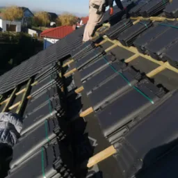 Pracownik w czapce i rękawicach montuje ciemne dachówki na przygotowanej konstrukcji, widok z góry na częściowo pokryty dach i okoliczne domy w tle.