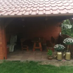 Drewniana altana z brązowym dachem krytym dach&oacute;wką ceramiczną, widoczne rynny, wewnątrz meble ogrodowe i dekoracje kwiatowe.