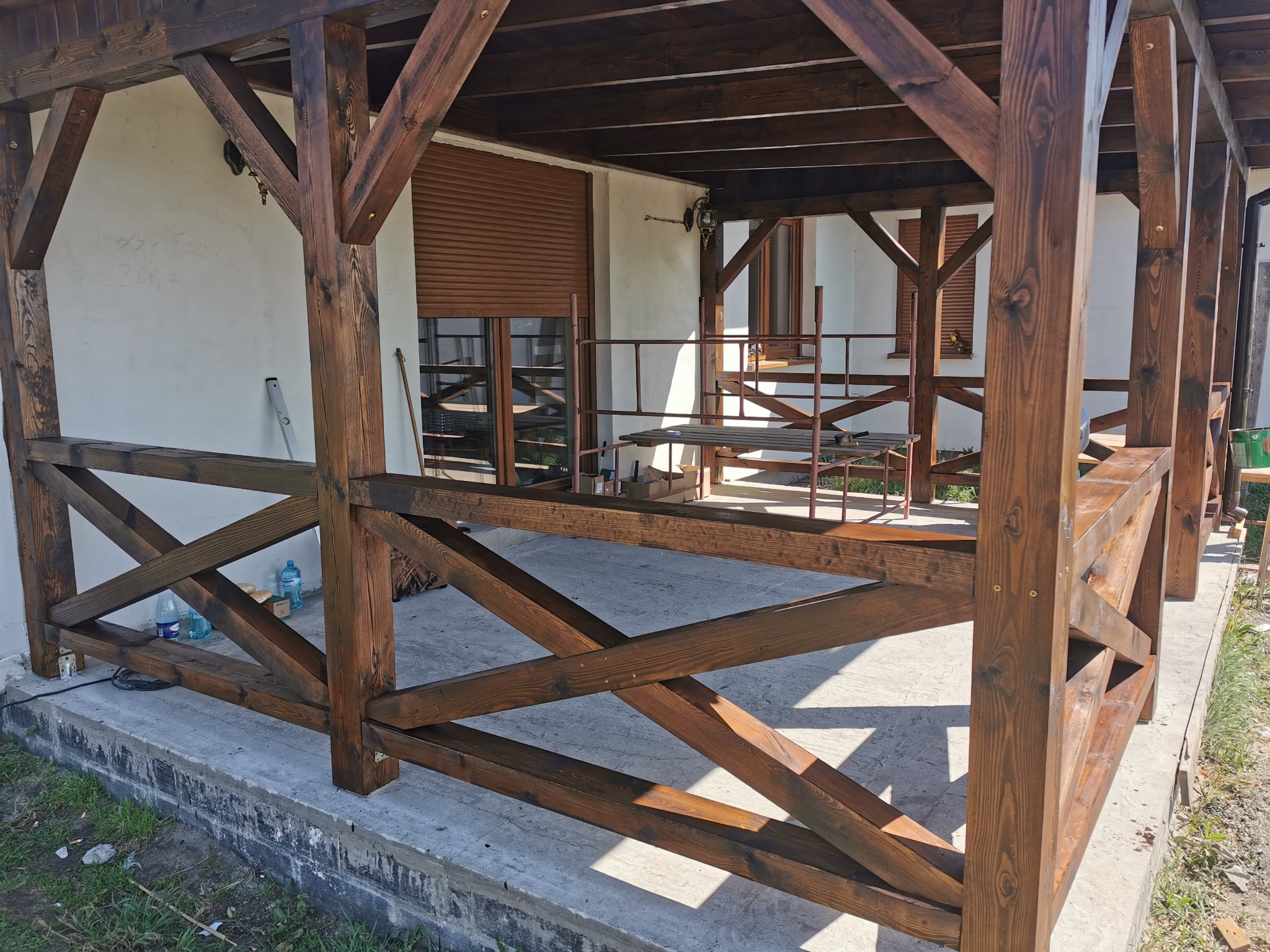 Drewniana konstrukcja z belek w kolorze ciemnego brązu, prawdopodobnie altana lub zadaszenie tarasu, z widoczną więźbą dachową i balustradą z krzyżujących się desek, na betonowej wylewce.