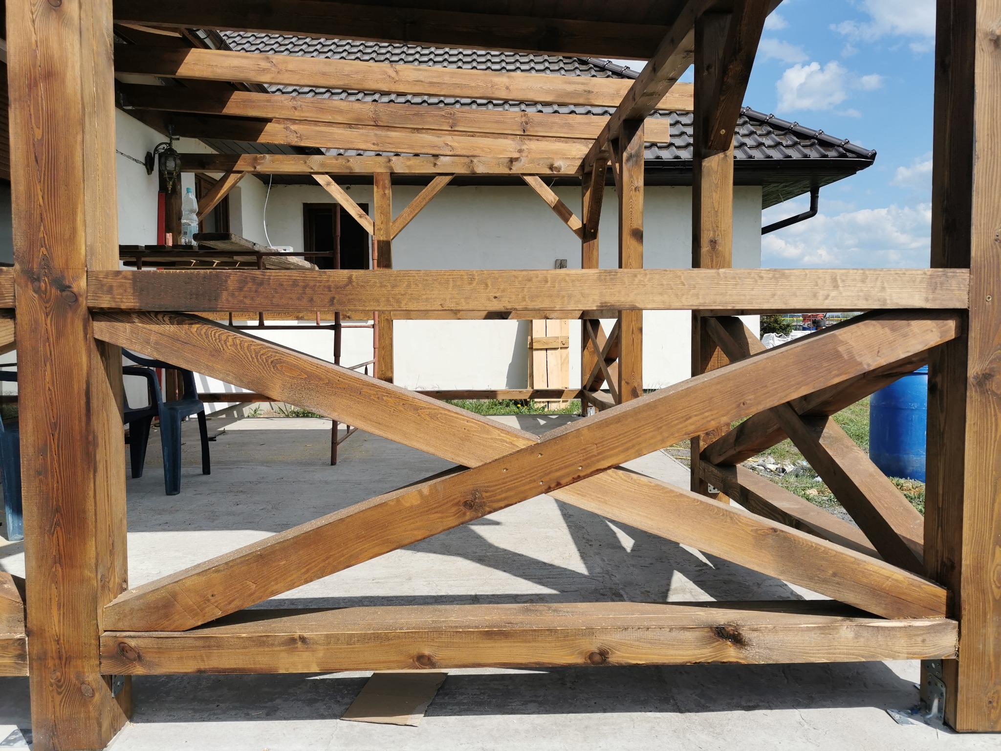 Drewniana konstrukcja altany w trakcie budowy, z widoczną więźbą dachową, fragmentem dachu pokrytego dachówką i błękitnym niebem w tle.