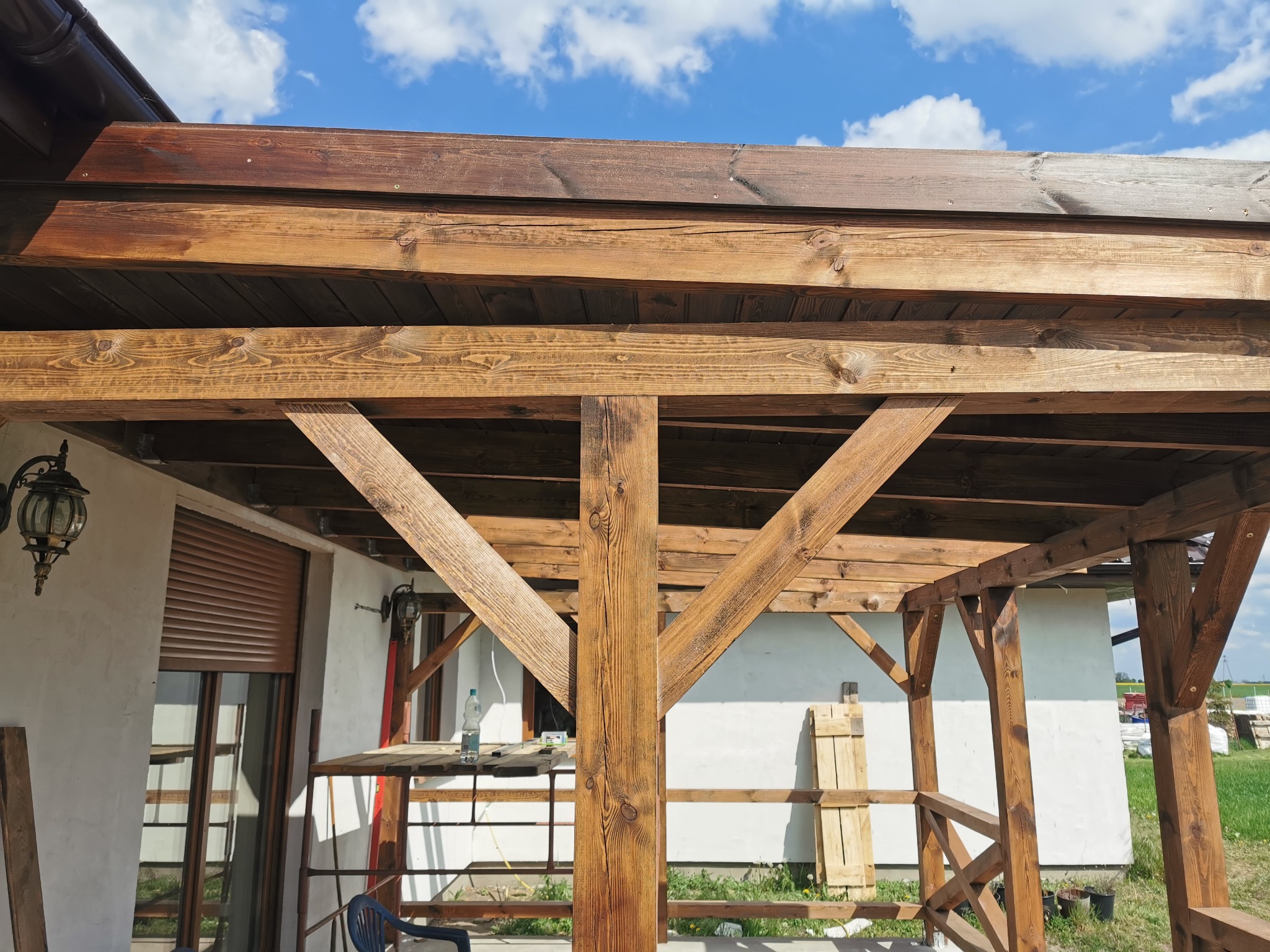 Drewniana konstrukcja z belek, prawdopodobnie zadaszenie tarasu lub werandy, z widoczną strukturą drewna i brązową impregnacją, na tle białej ściany budynku i błękitnego nieba.