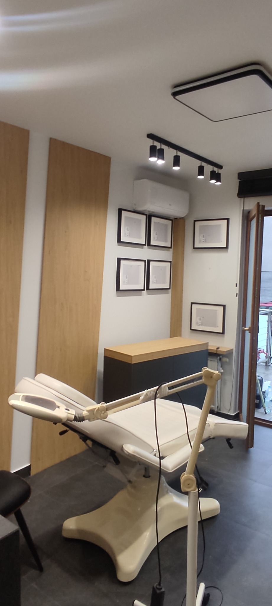 Nowoczesne wnętrze gabinetu kosmetycznego z białym fotelem zabiegowym z regulowanym oświetleniem, drewnianą zabudową ściany i minimalistycznymi obrazkami w czarnych ramkach