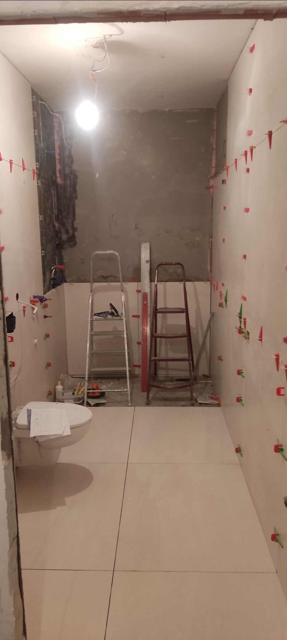 Remont łazienki w trakcie: ułożone jasne płytki podłogowe, ściany przygotowane do położenia płytek z użyciem klipsów poziomujących, dwie drabiny ustawione w pomieszczeniu, widoczne narzędzia...