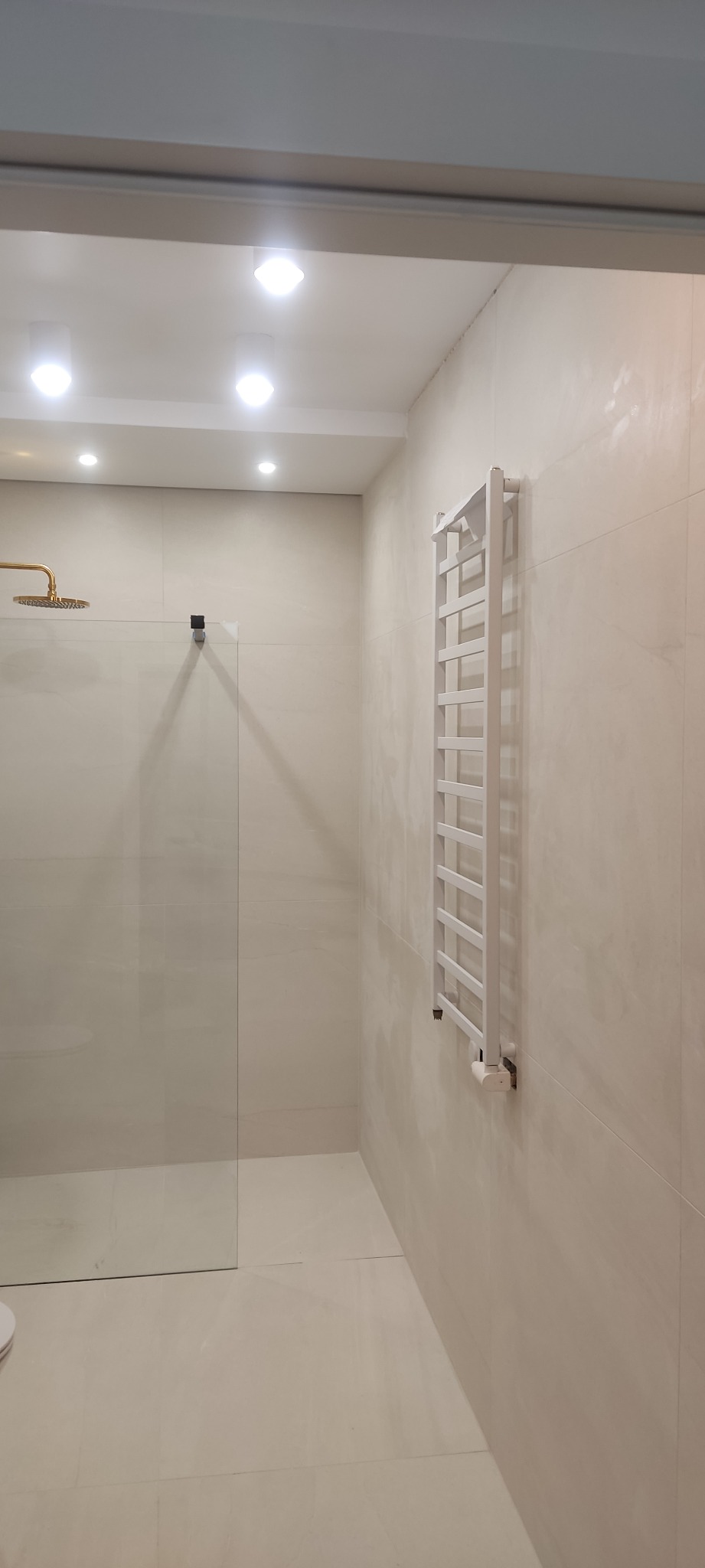 Nowoczesna łazienka z kabiną prysznicową ze złotą deszczownicą, grzejnikiem drabinkowym i beżowymi płytkami wielkoformatowymi.